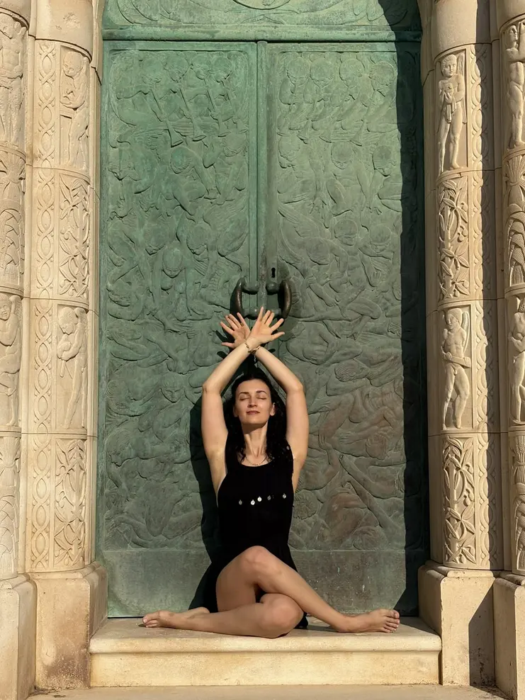 Sonia sitting in a yoga pose with her arms raised, in front of tall green bronze temple doors.